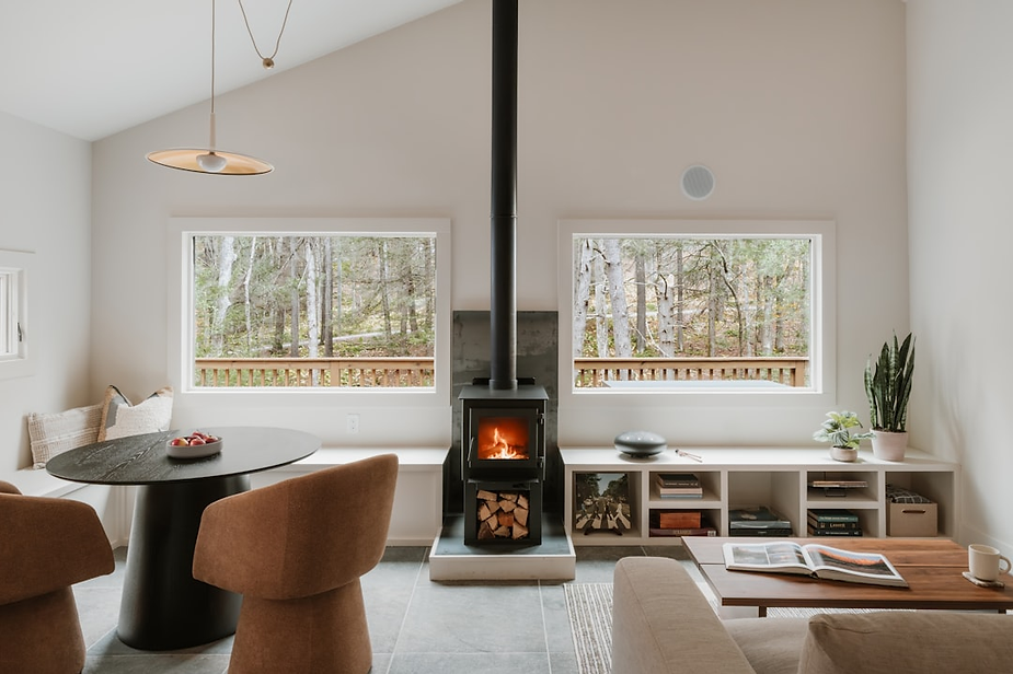 Cozy living room featuring a modern wood stove, circular dining table with fruit, and large windows overlooking a forest, emphasizing warmth and comfort for winter in Eatonville.