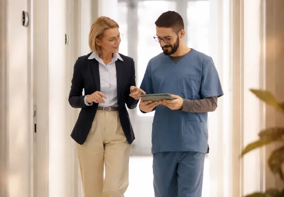 Healthcare professional in blue scrubs showing a tablet to a an infusion center executive while walking in a hallway.