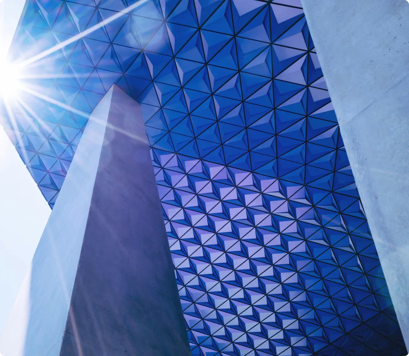 Low-angle view of geometric blue glass ceiling with triangular patterns supported by large concrete pillars, sun flare on the left.
