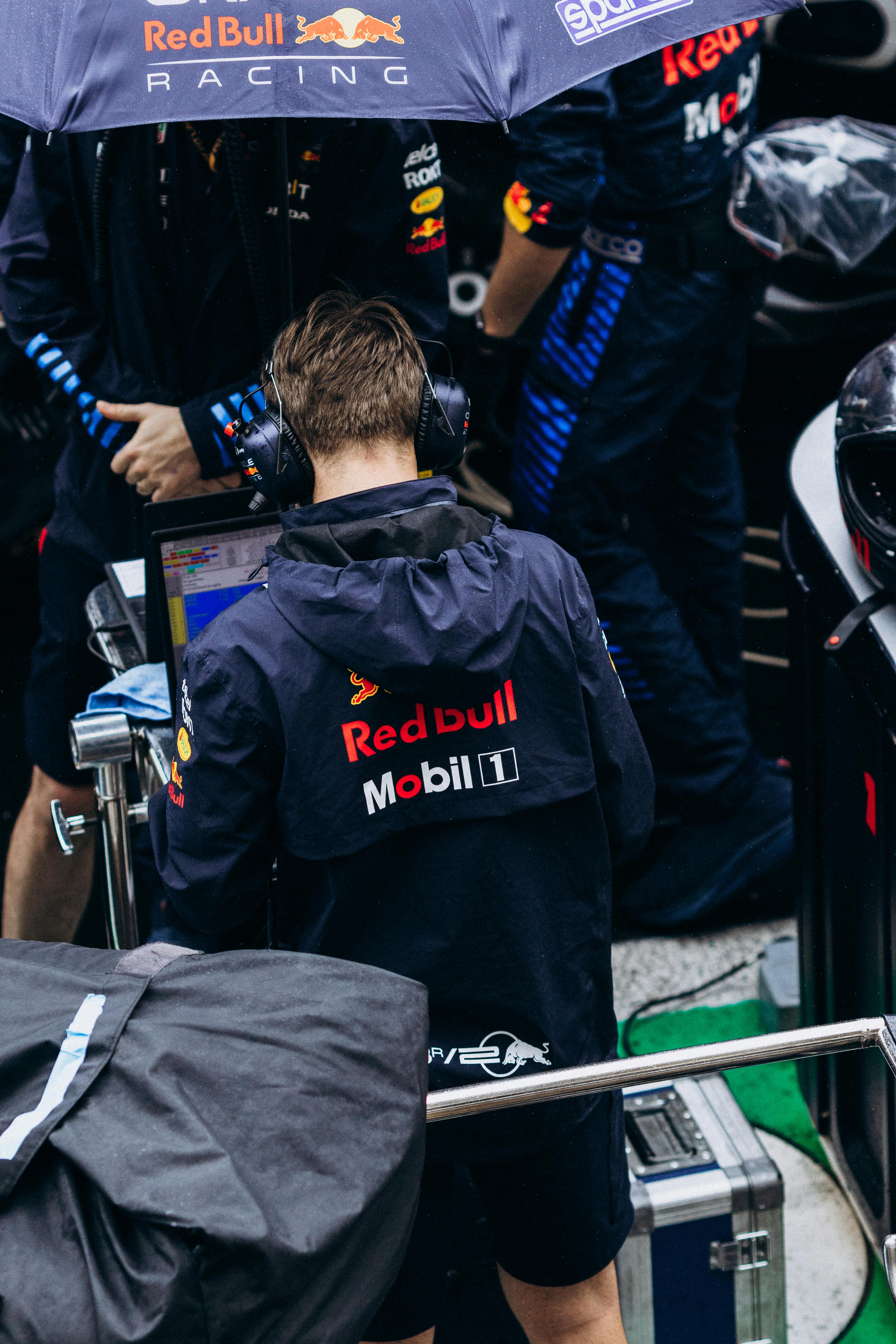 Person in Red Bull Racing jacket and headset working on laptop under umbrella.