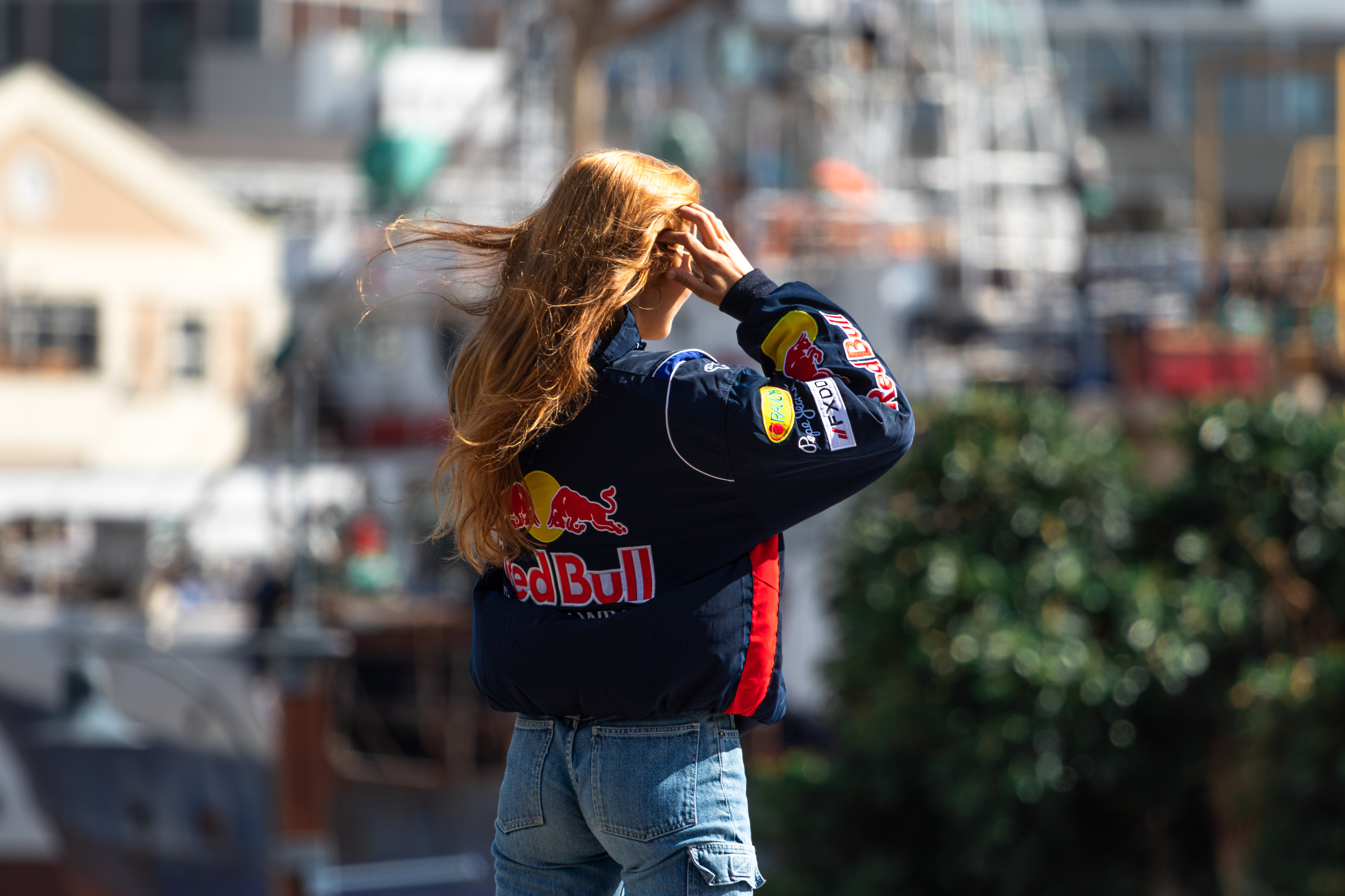 Person with long hair wearing a Red Bull jacket and jeans, standing outdoors with blurred background.
