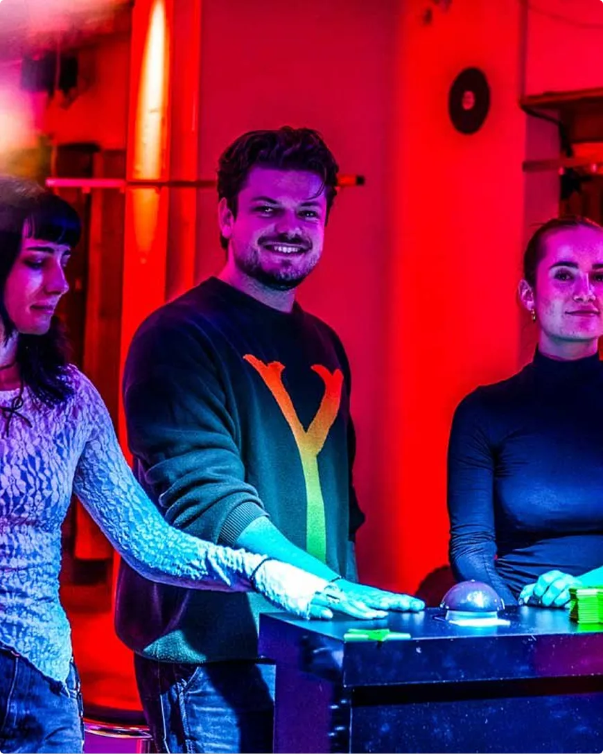 Three people standing by a table with a button in a room with red and pink lighting, one smiling man in the center wearing a black sweater with a colorful Y design.