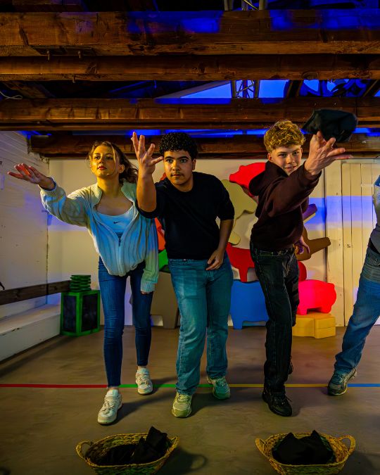 Five people standing at illuminated podiums in a colorful, neon-lit room, possibly playing a quiz or game show.