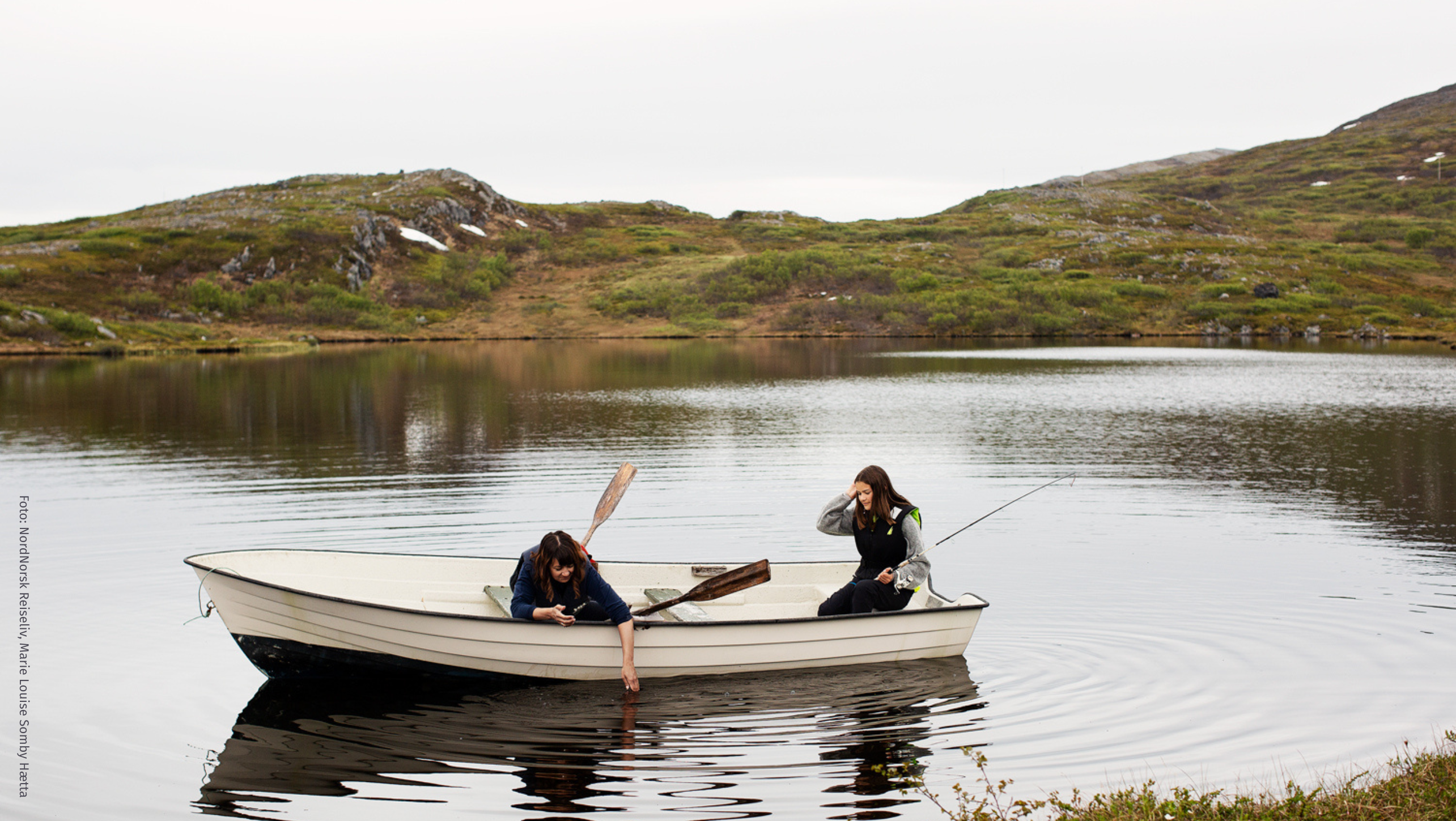 Tre mennesker koser seg i fjæra med fjorden i bakgrunnen. De har tent bål og spiser mant.