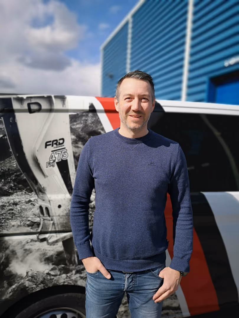 Smiling man in blue sweater standing outdoors in front of a vehicle with graphics and a blue building behind.