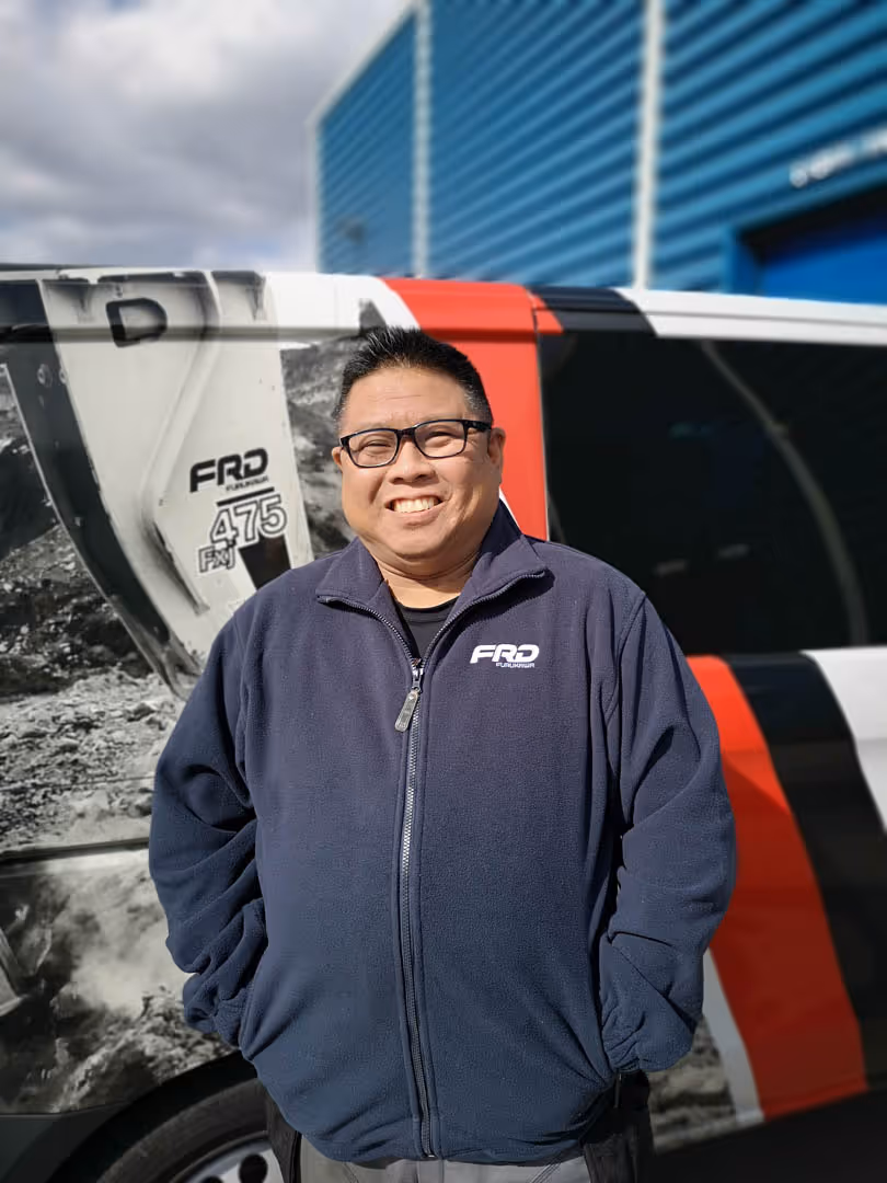 Smiling man wearing glasses and a navy FRD zip-up jacket stands in front of a branded vehicle.