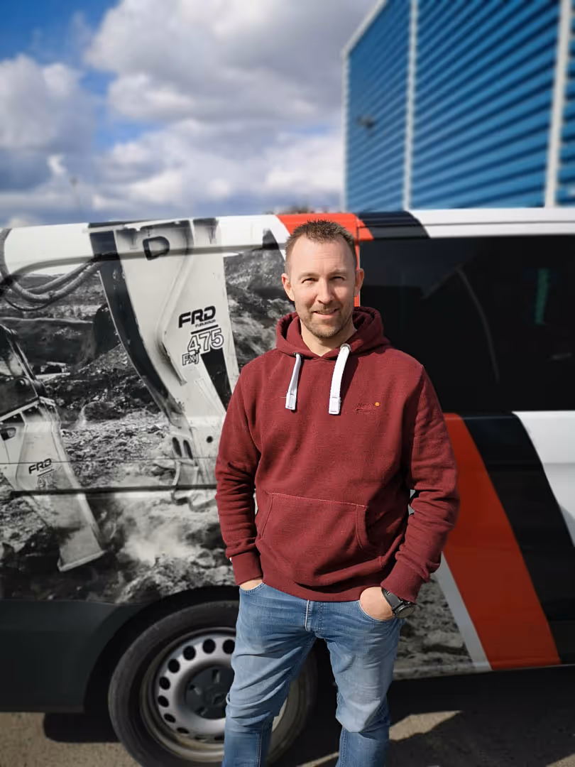 Man in a maroon hoodie and jeans stands smiling with hands in pockets by a van with machinery graphics.