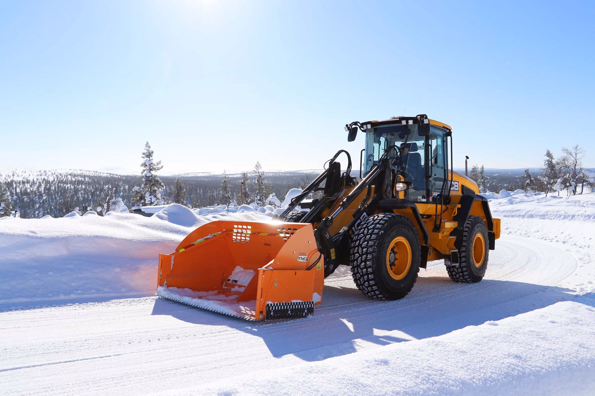 Gul hjullaster med oransje snøfreser rydder en snødekt vei i vinterlandskap.