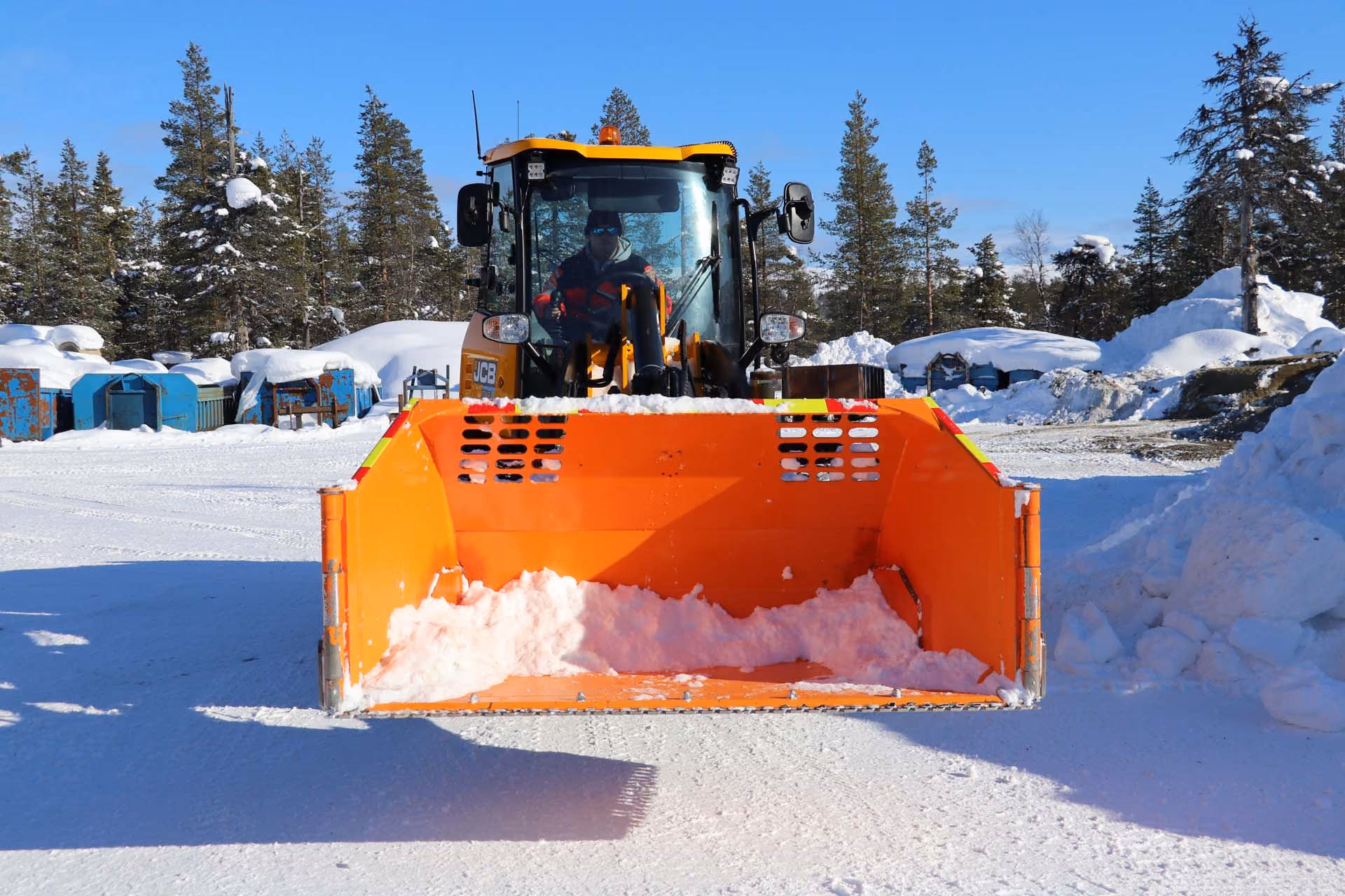 Hjullaster med oransje skuff foran fylt med snø og skog i bakgrunnen.