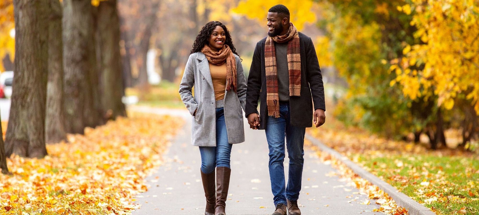 Two people walking along tree-lined street with fallen leaves.