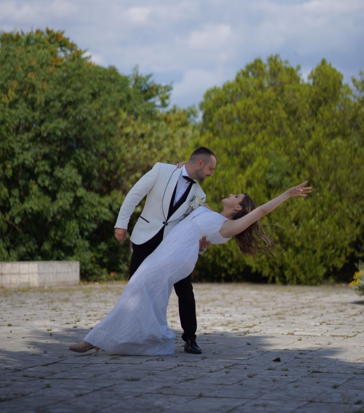 Bride in a white dress leaning backward in a dance dip held by groom in a white jacket and black pants outdoors. Bojan Mitevski wedding
