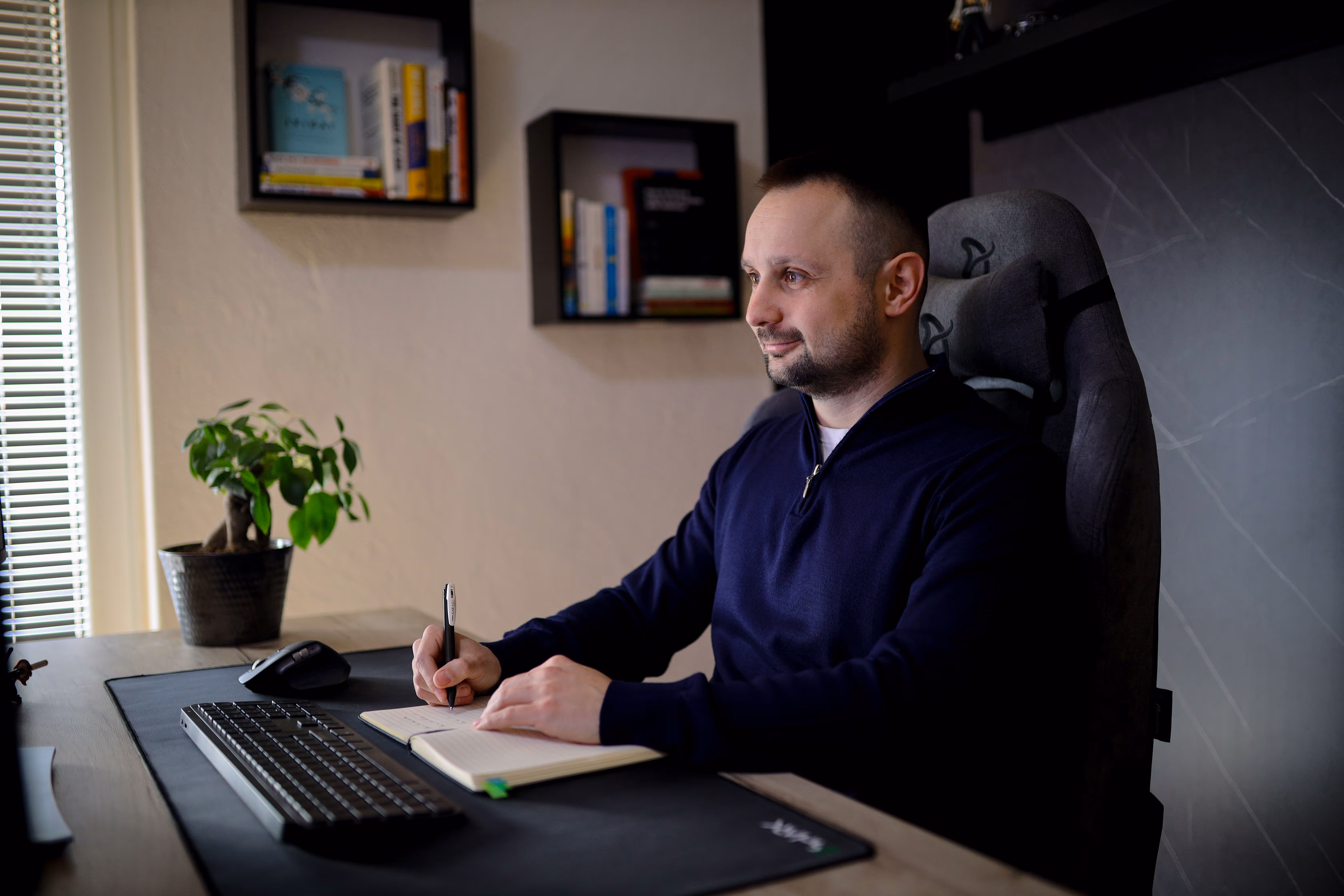Bojan Mitevski at a desk writing in a notebook with a computer keyboard and mouse nearby, shelves with books on the wall.