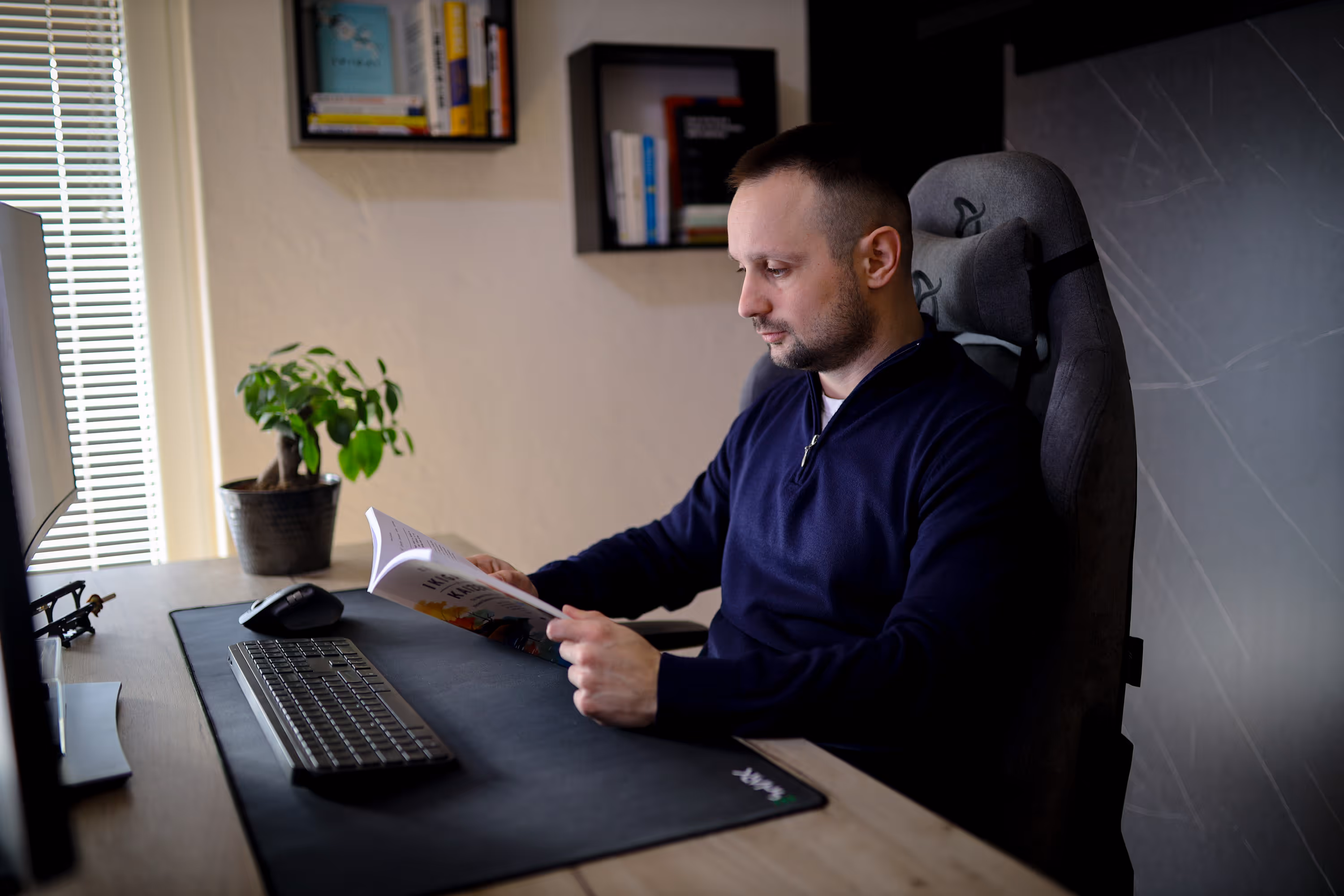 Man in a navy sweater sitting at a desk reading a book in a modern office.