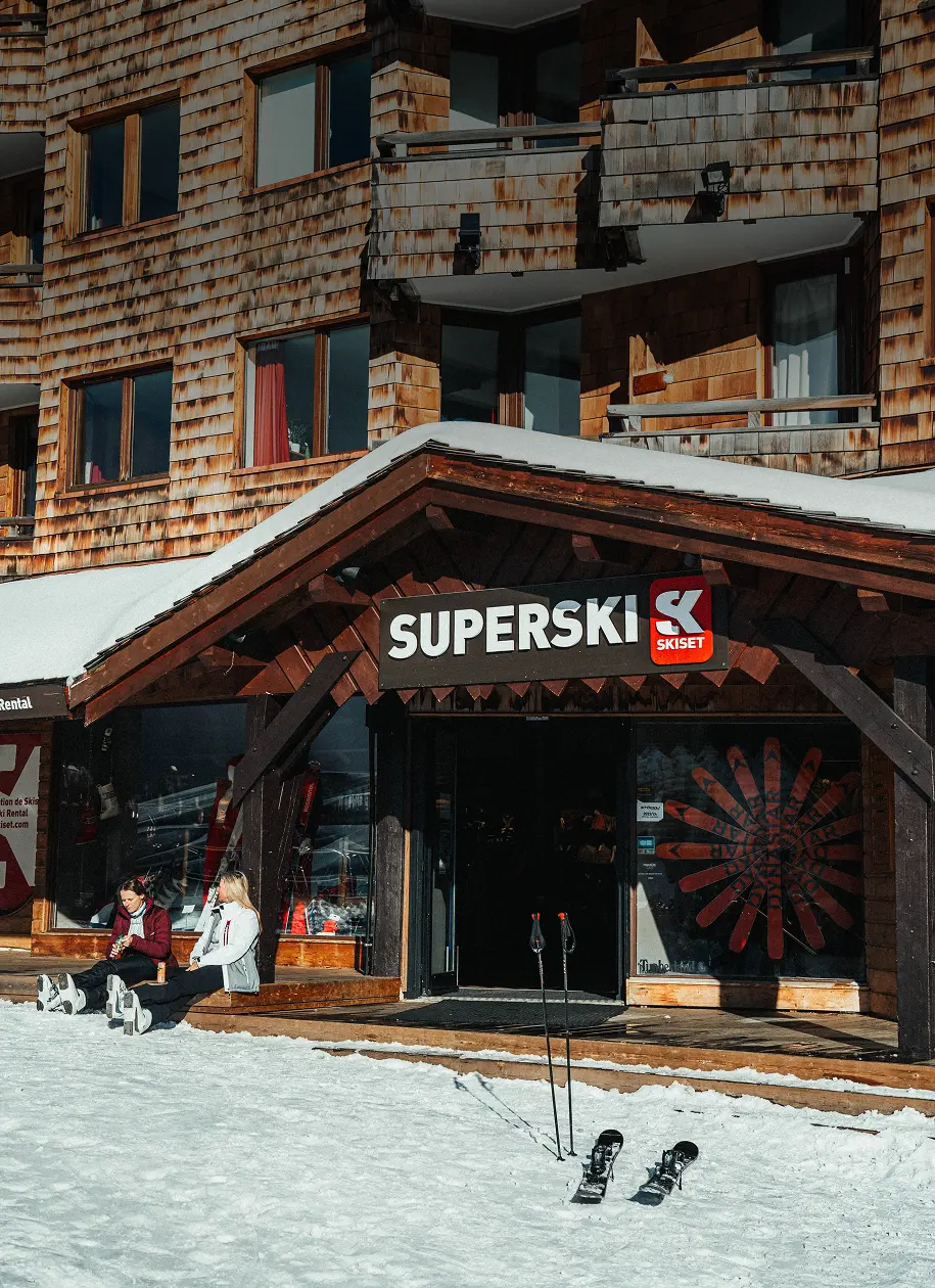 Deux personnes assises sur un banc enneigé devant un magasin SUPERSKI en bois avec skis posés dans la neige.