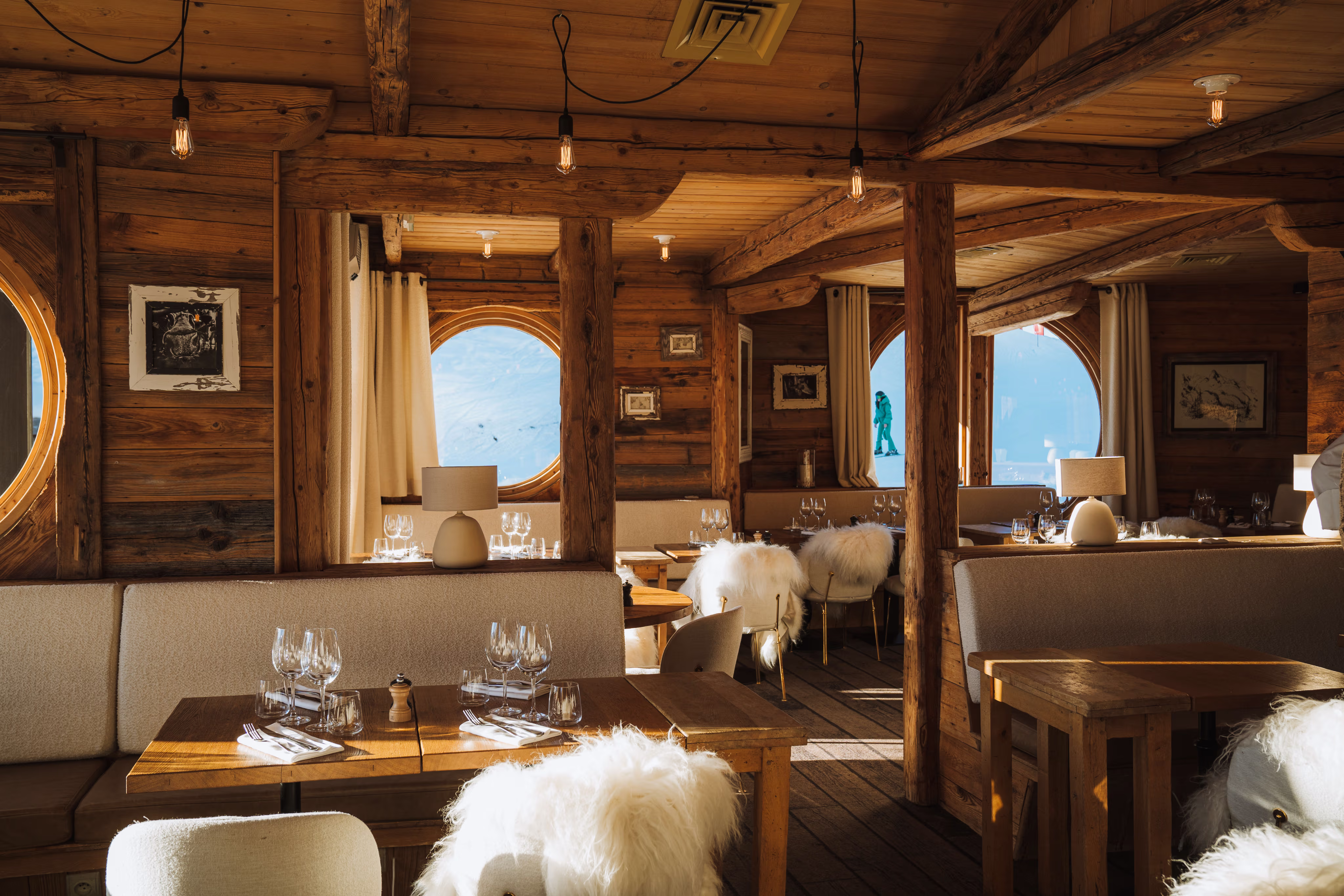 Intérieur de restaurant en bois avec tables dressées et vue sur un skieur dehors par fenêtres rondes.
