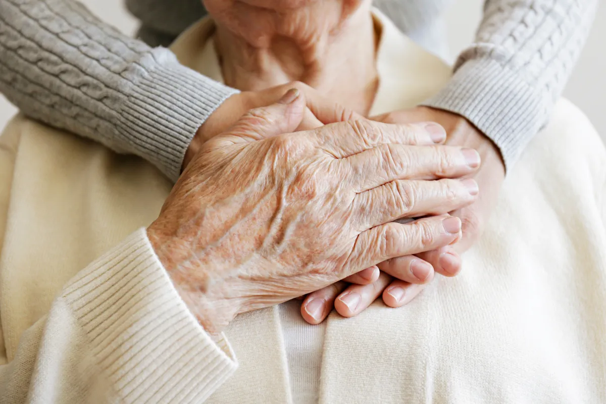 Caregiver’s hands gently holding elderly woman’s hands over her chest in a comforting gesture.