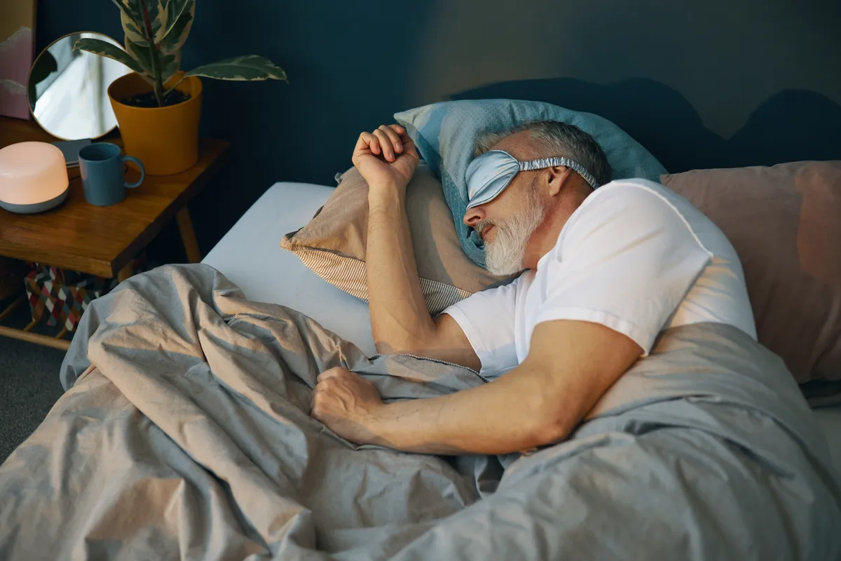 Older man sleeping on his side in bed with a blue eye mask and white t-shirt.