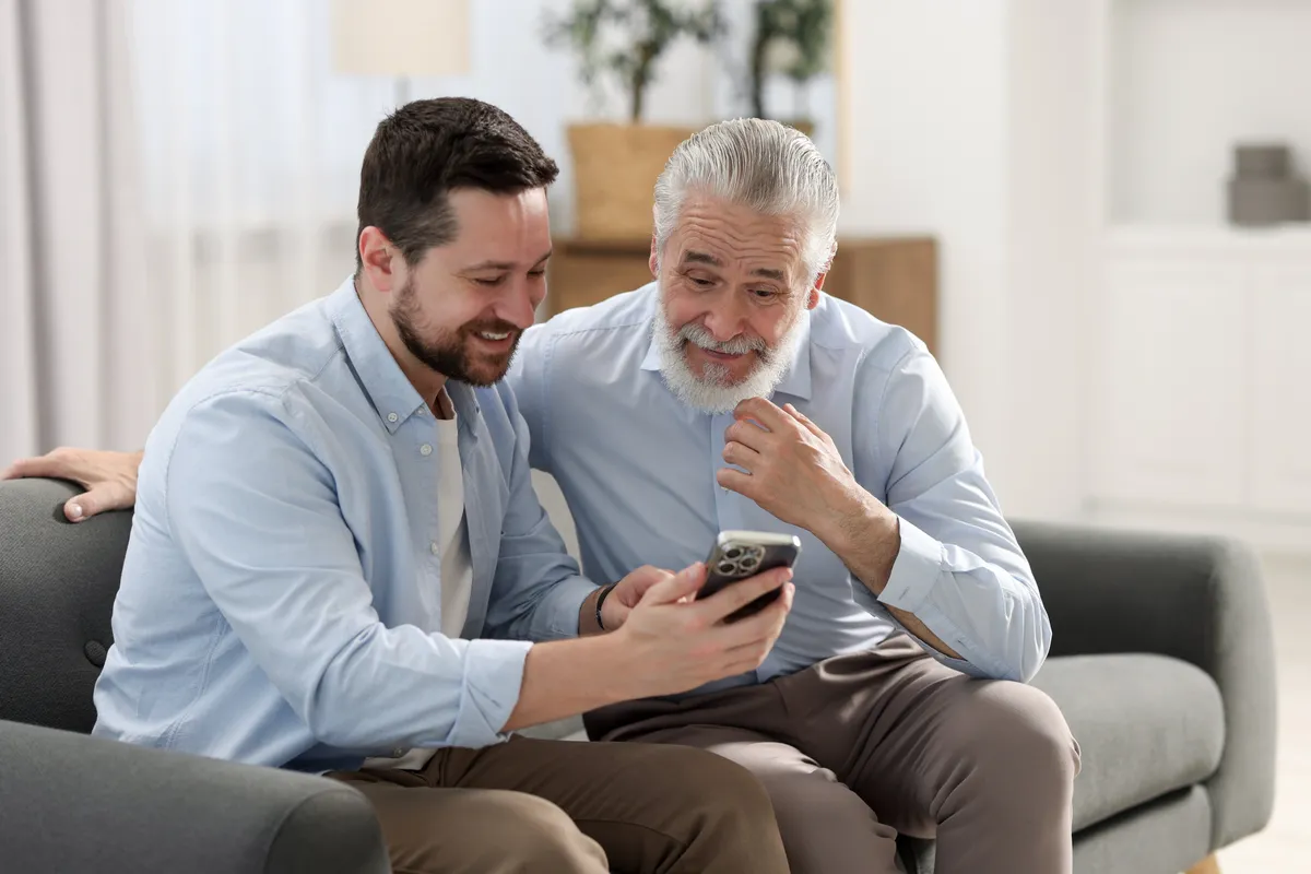 Smiling adult son and senior father sitting on a couch looking at a smartphone together.