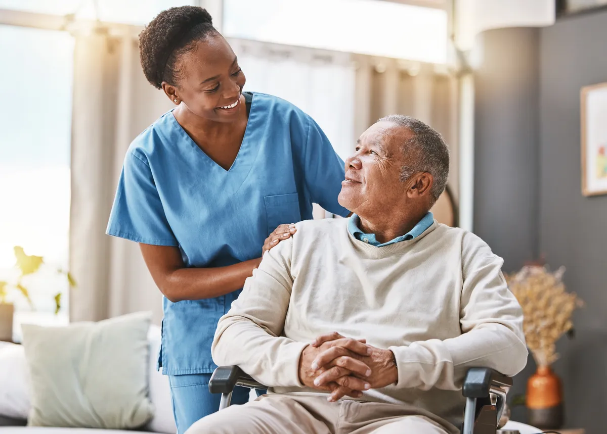 Smiling home caregiver in blue scrubs standing behind a senior man sitting in a wheelchair.