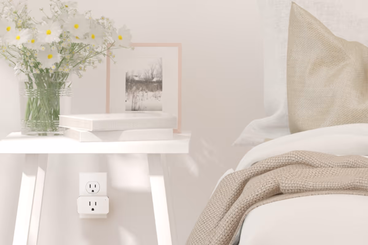 Small white electronic device plugged into a wall outlet beneath a white side table with flowers and books next to a bed.