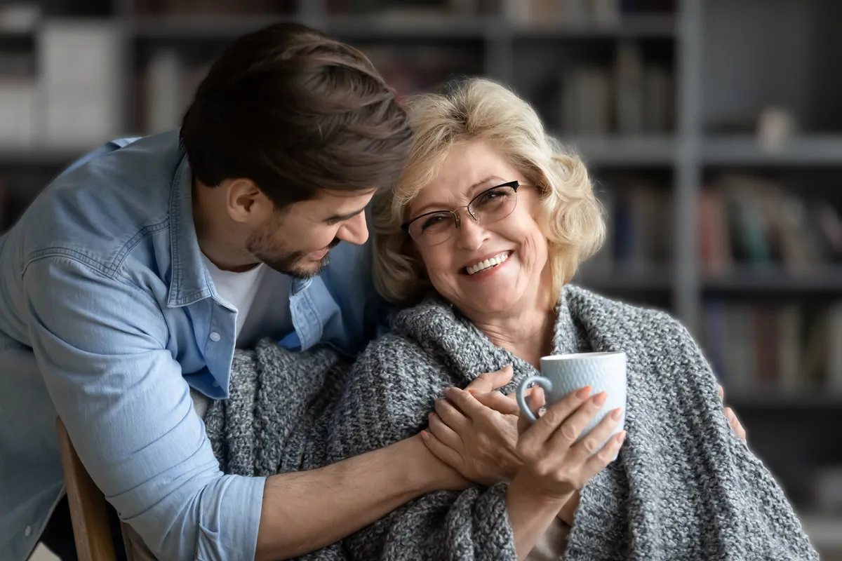 Adult son wrapping blanket around smiling senior mother.