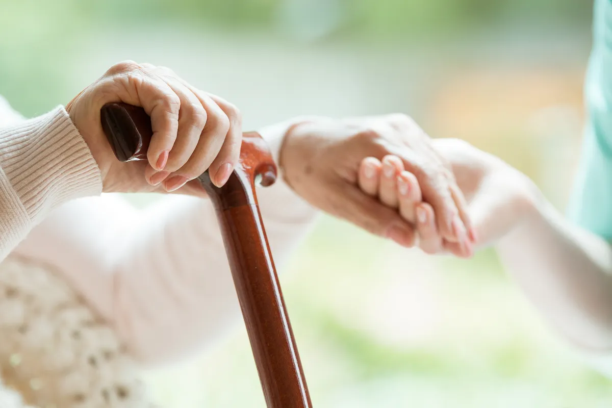Elderly hands on walking cane with caregiver support.