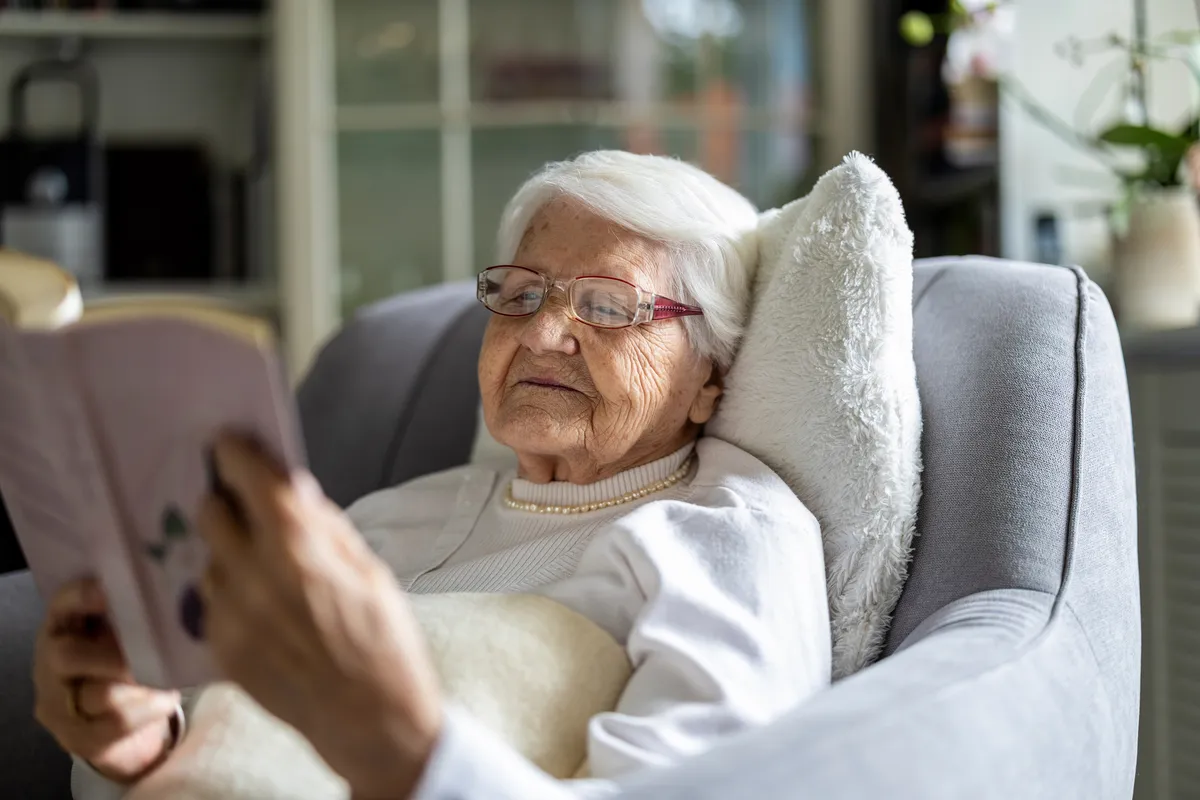Elderly woman reading book in armchair at home.