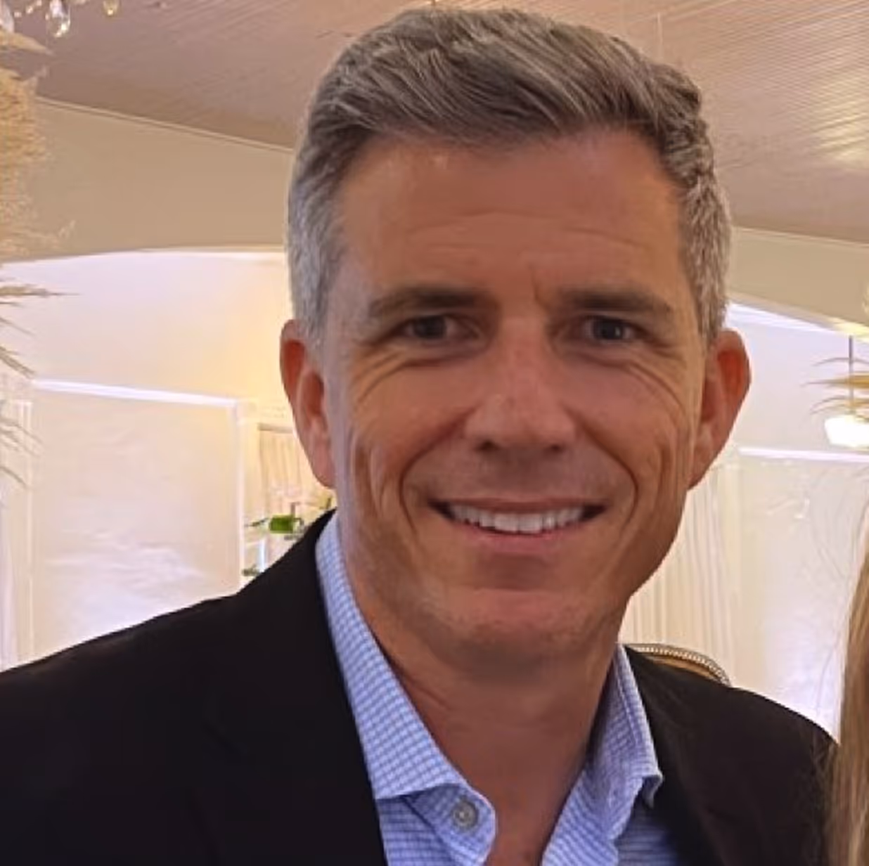 Portrait of a young man with blue eyes and short blonde hair wearing a blue suit jacket and white shirt against a dark background.