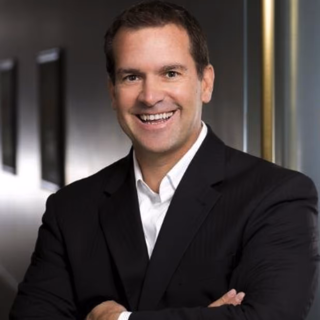Portrait of a young man with blue eyes and short blonde hair wearing a blue suit jacket and white shirt against a dark background.