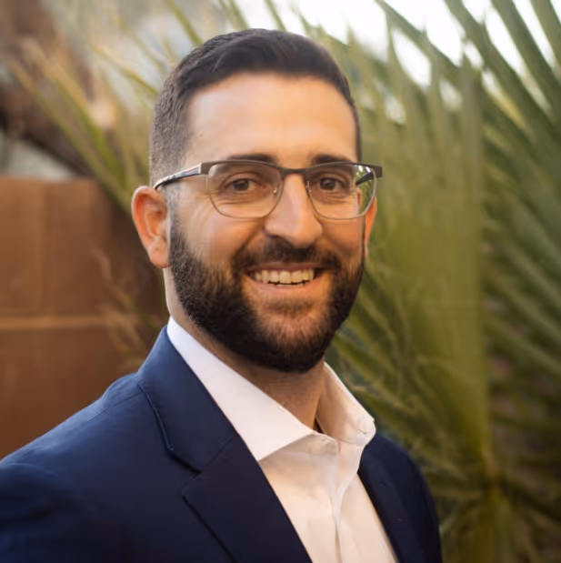 Portrait of a young man with blue eyes and short blonde hair wearing a blue suit jacket and white shirt against a dark background.
