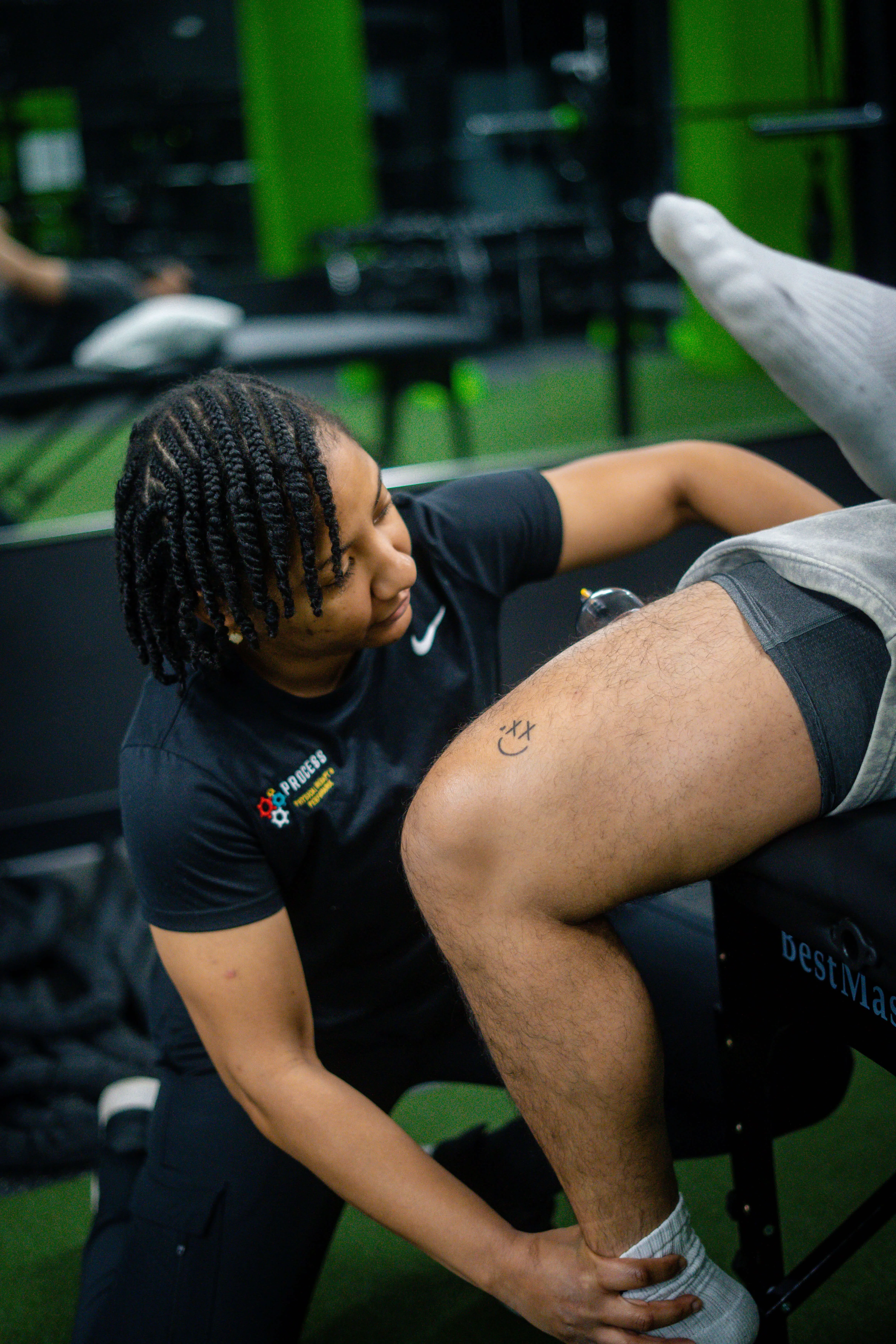 A physical therapist with braided hair assists a person lying on a therapy table by holding their ankle during a leg exercise in a gym.