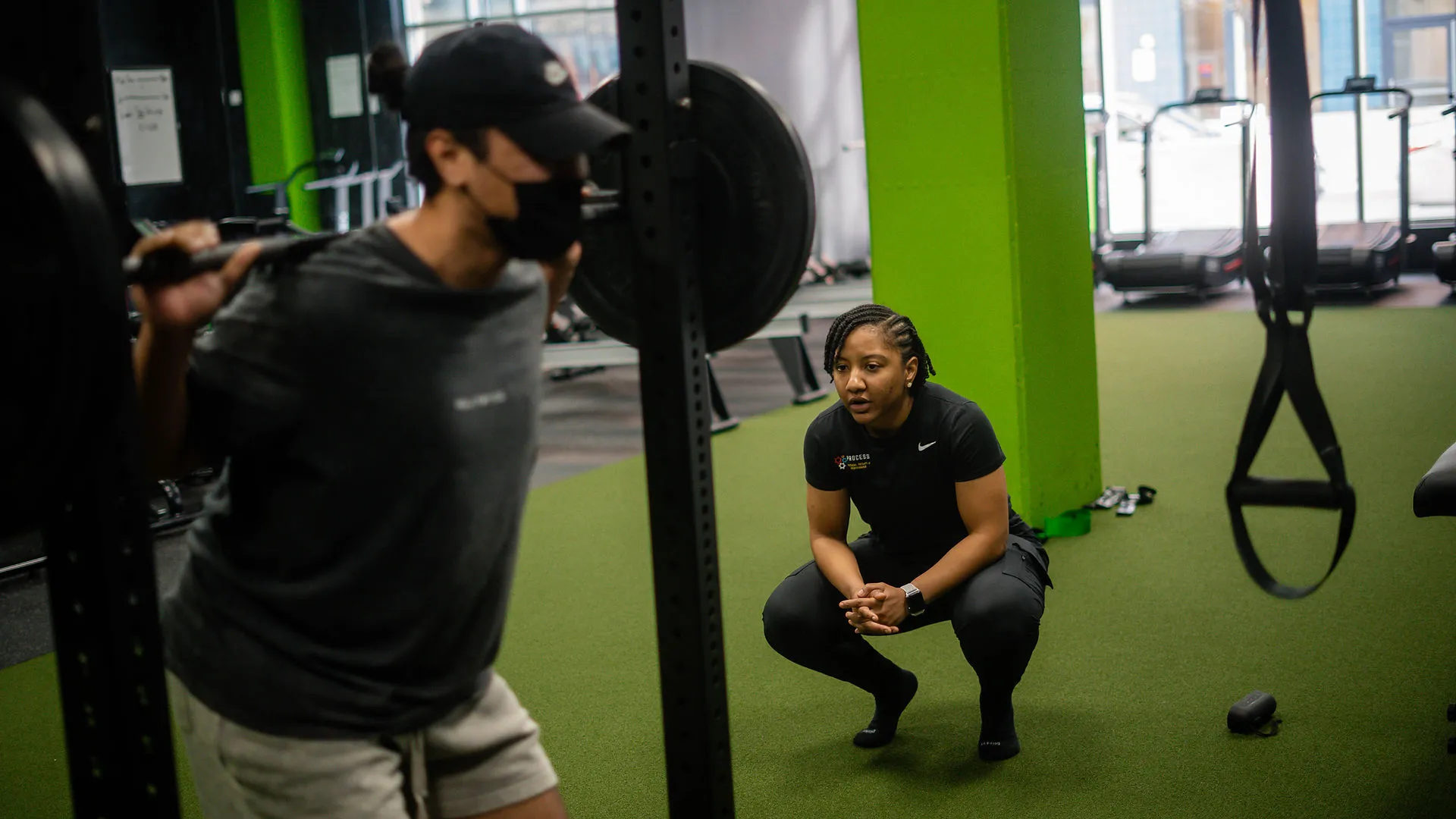 Woman trainer squatting on green turf floor coaching a man lifting a barbell with weights in a gym.