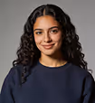 Portrait of a smiling young woman with curly dark hair wearing a navy blue sweater against a gray background.