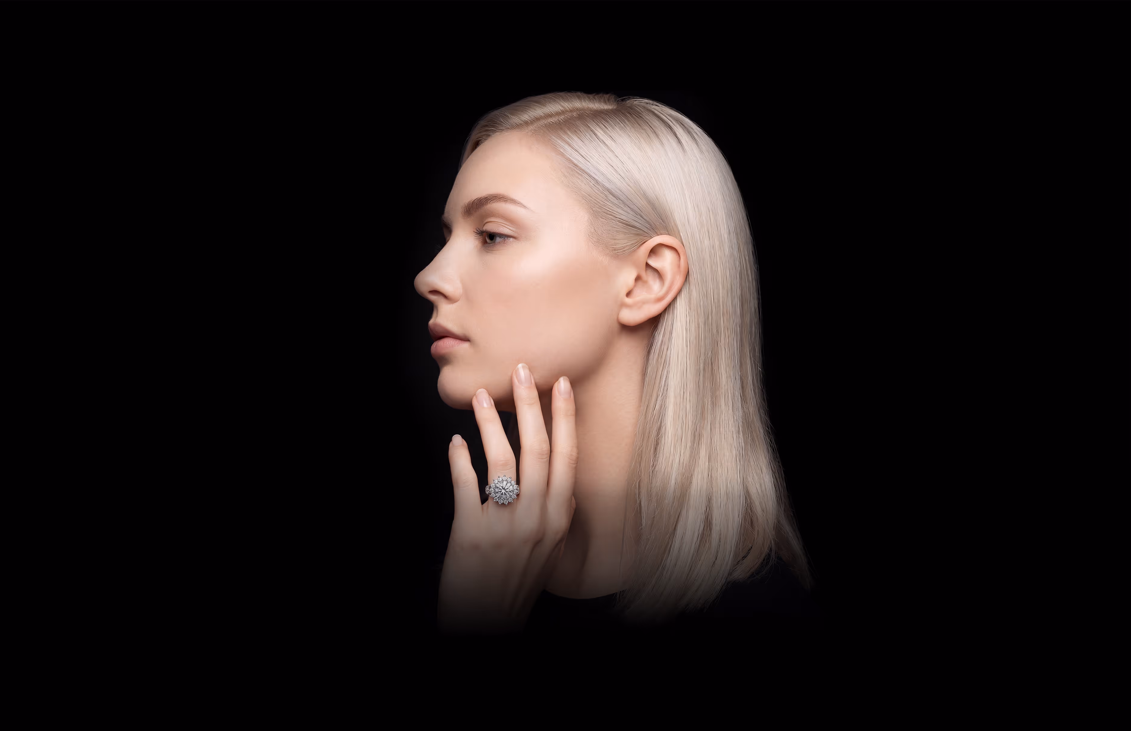 Profile of a blonde woman with straight hair showing a large diamond ring on her finger against black background.