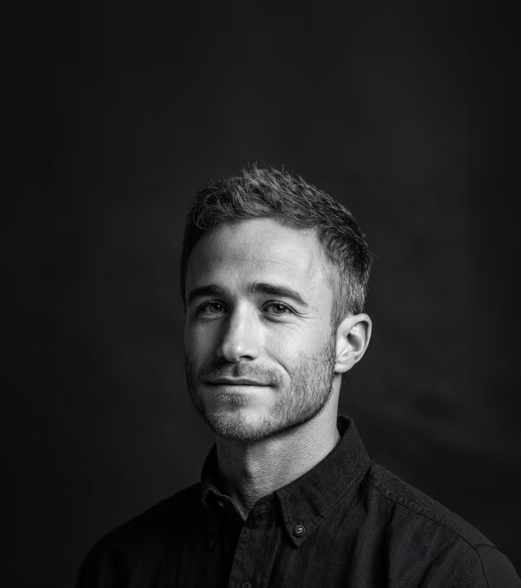 Black and white portrait of a man with short hair and stubble wearing a buttoned shirt against a dark background.