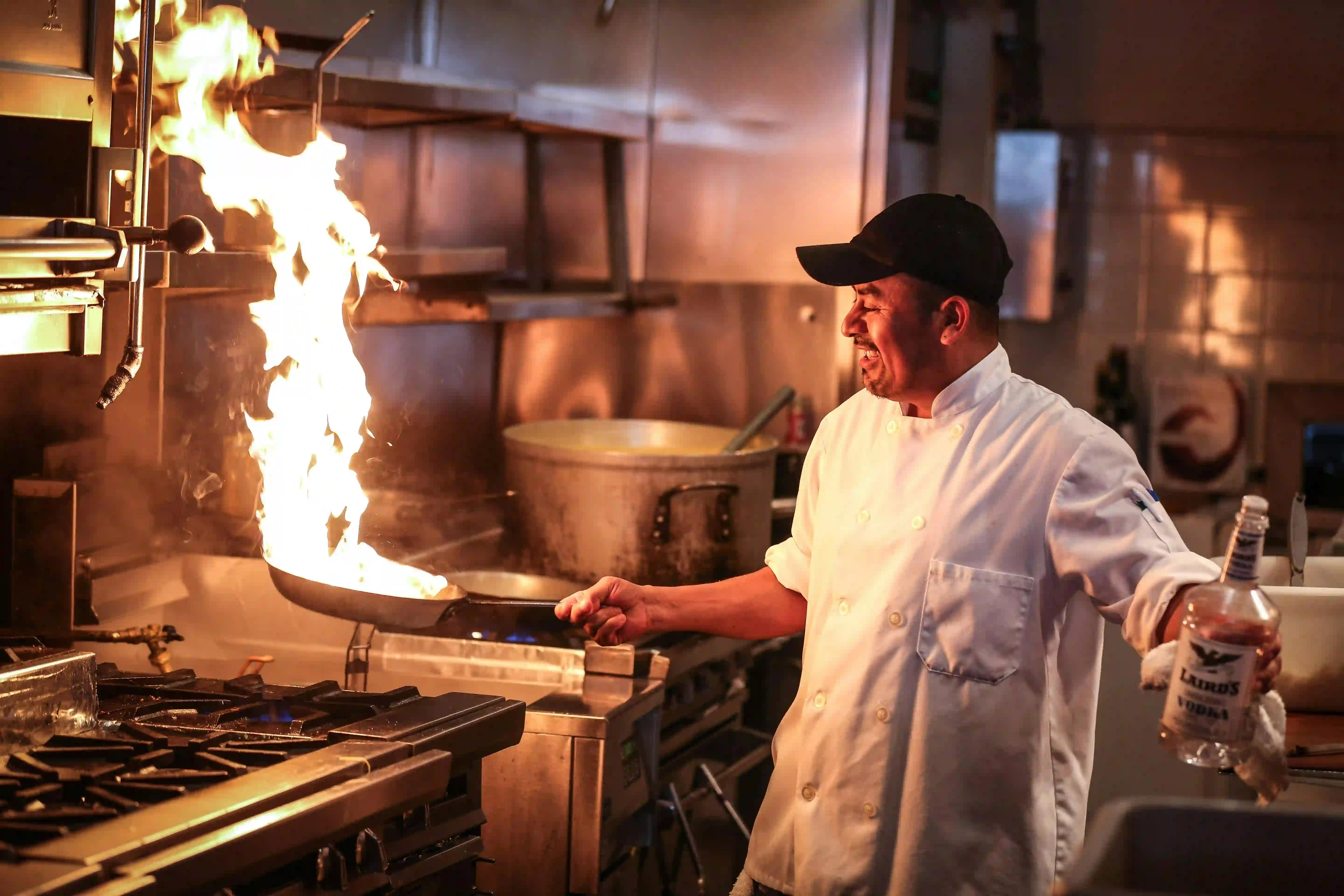 Chef souriant en toque noire flambant une poêle dans une cuisine professionnelle tout en tenant une bouteille de vodka.