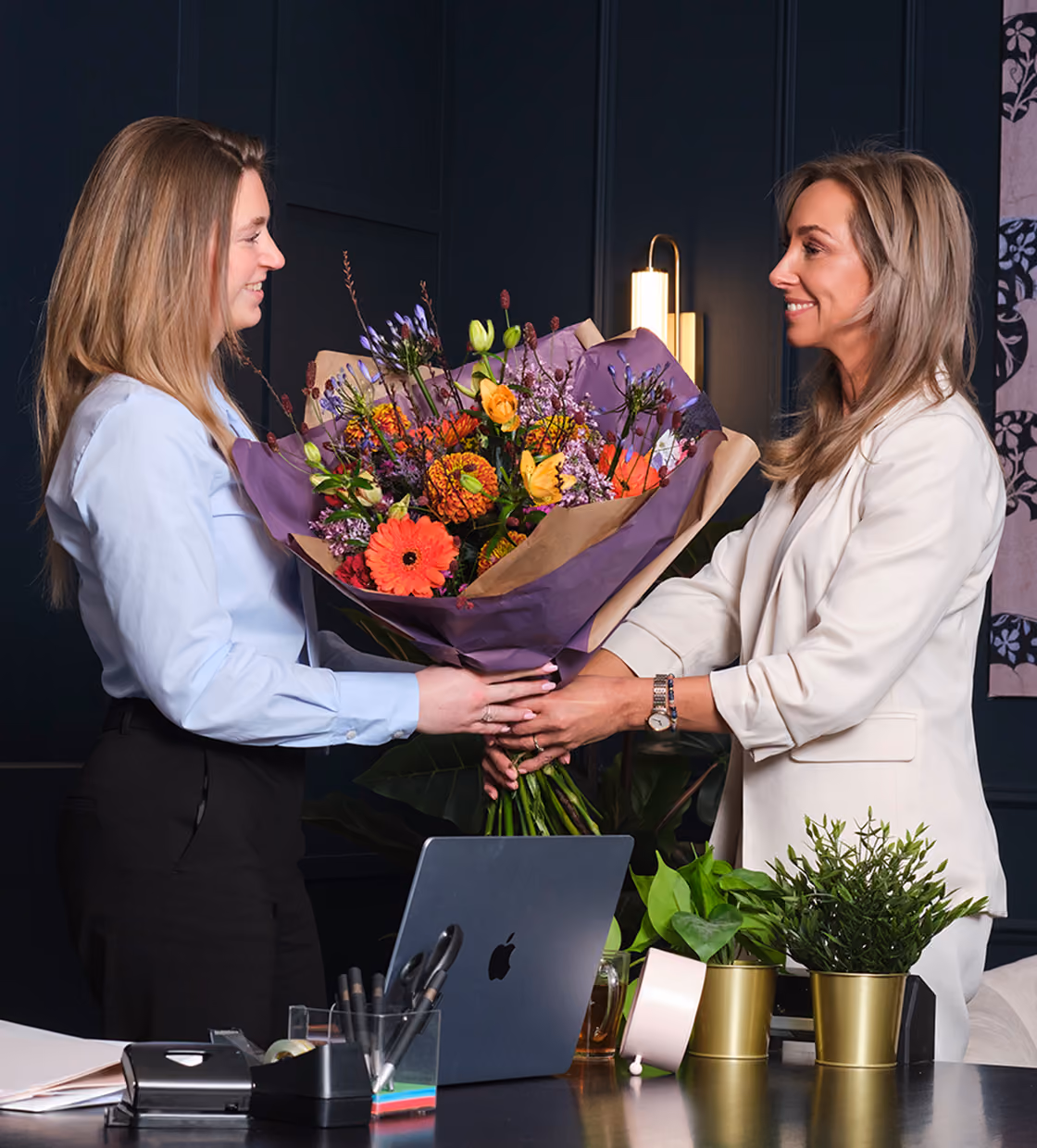 Twee vrouwelijke collega's wisselen een kleurrijk boeket bloemen uit op kantoor.