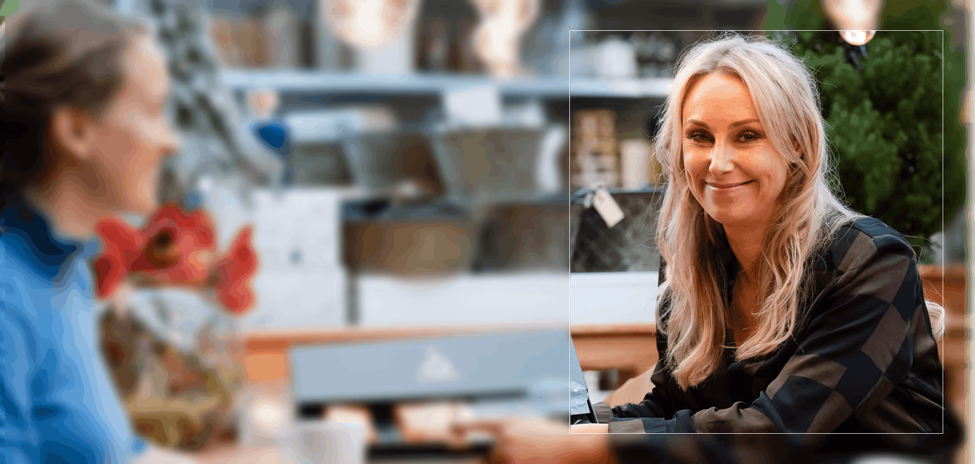 Vrouw met blond haar en geruit shirt glimlacht terwijl ze aan een tafel zit in een café.