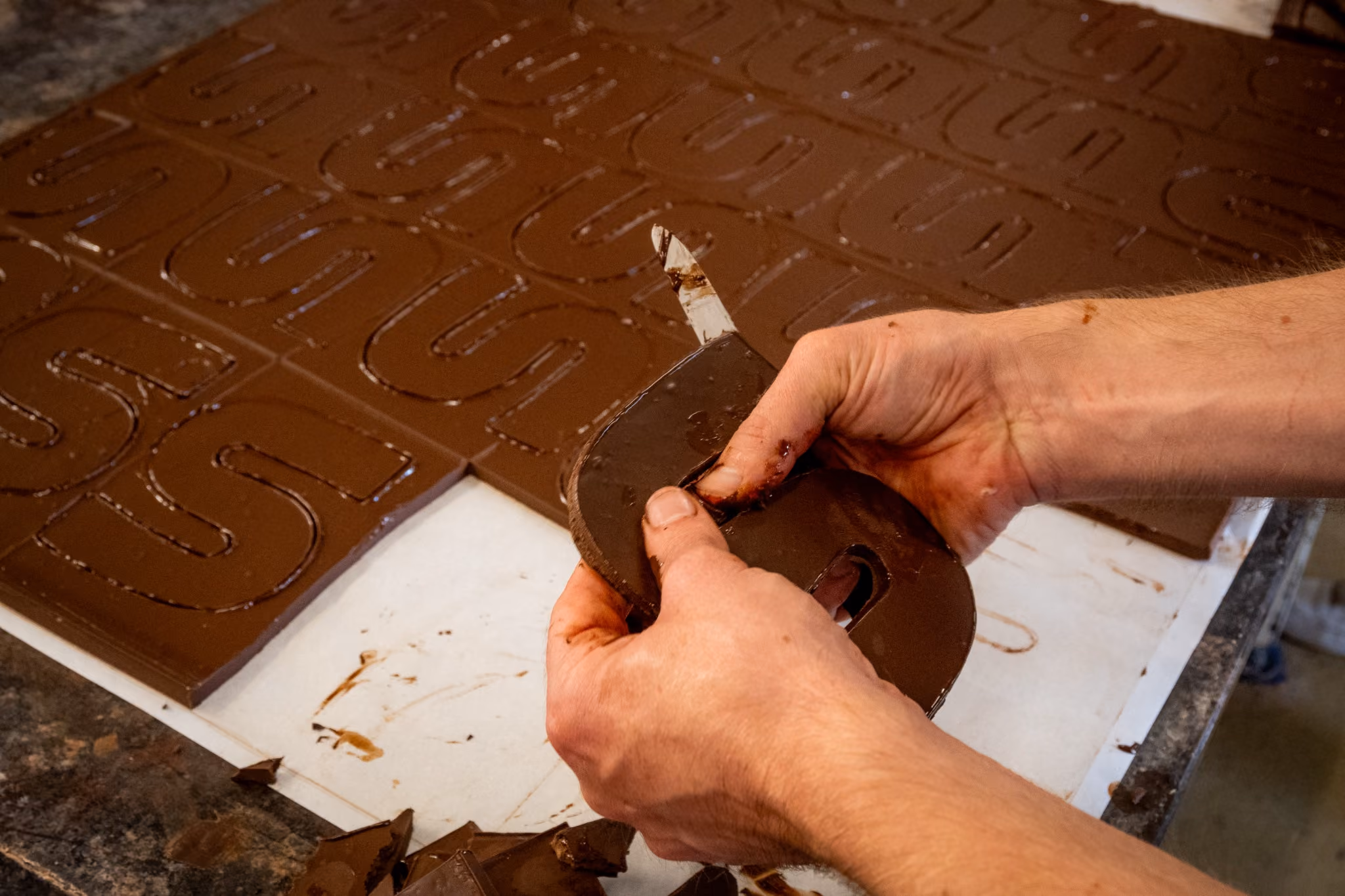 Iemands handen breken grote chocoladeletters in stukken op een werkblad.