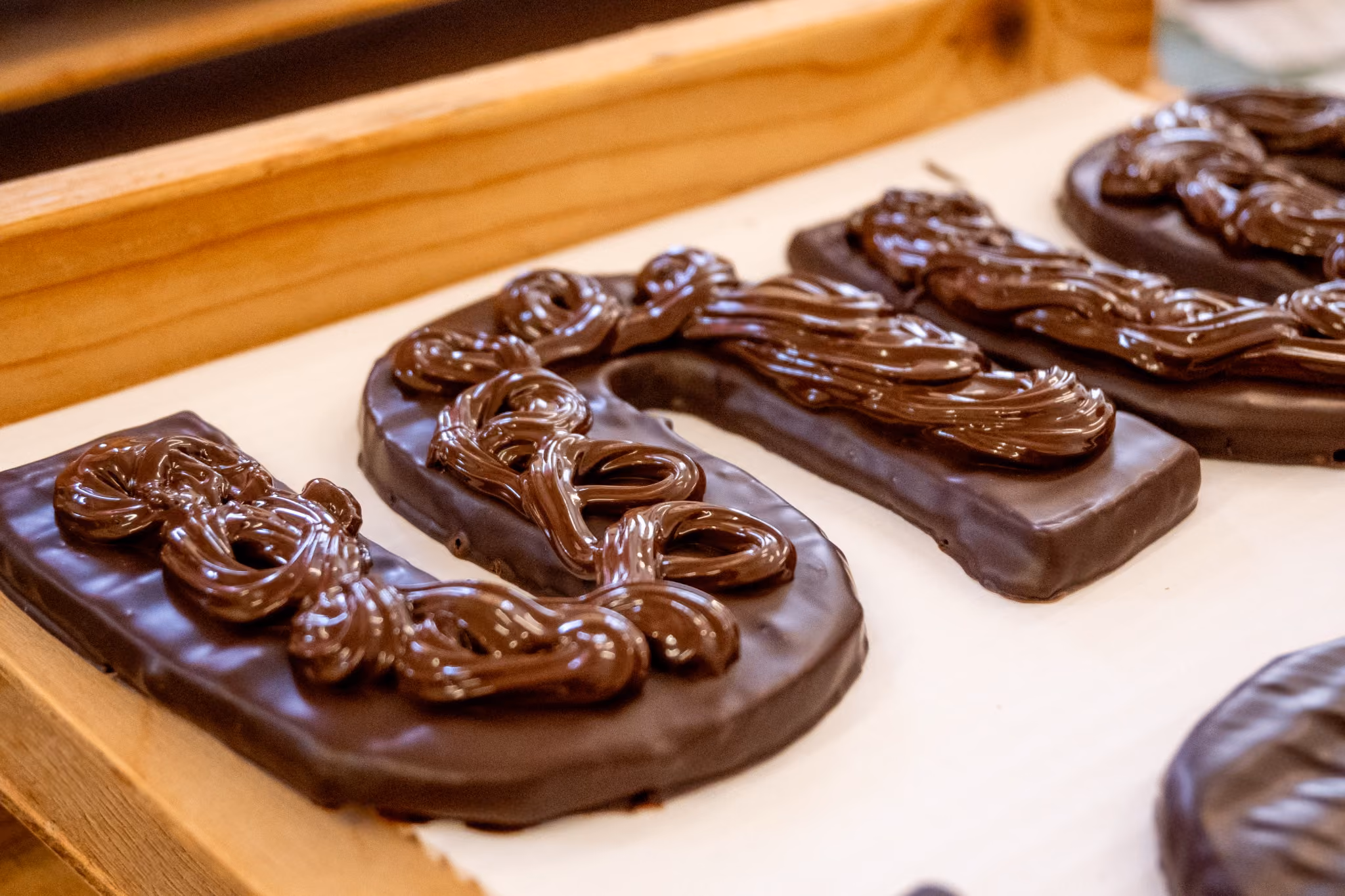 Vier chocoladeletters met glanzende chocoladedecoratie op bakpapier in houten bak.