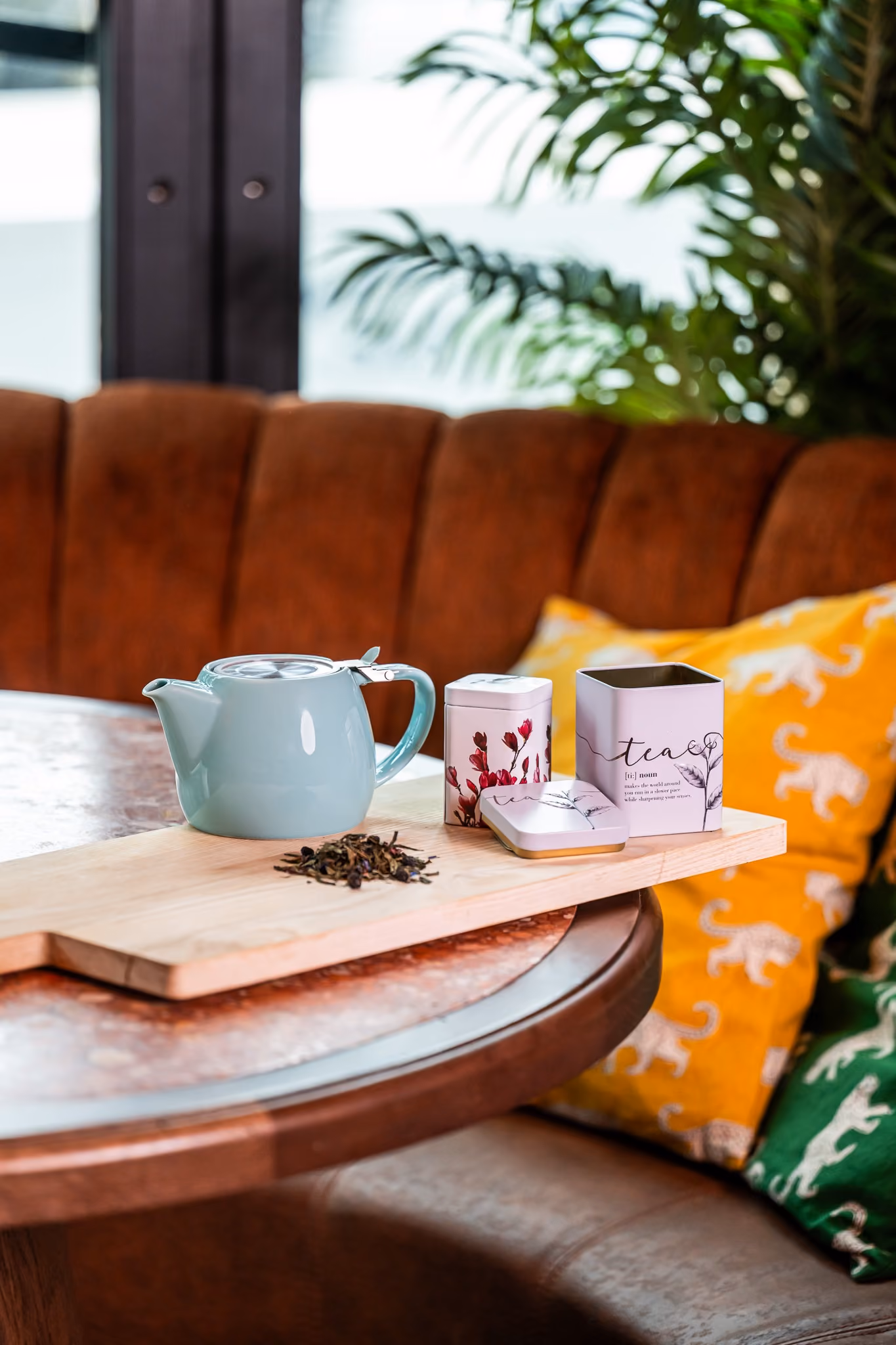 Theepot en drie theedozen op een houten plank op een tafel, met kussens en planten op de achtergrond.
