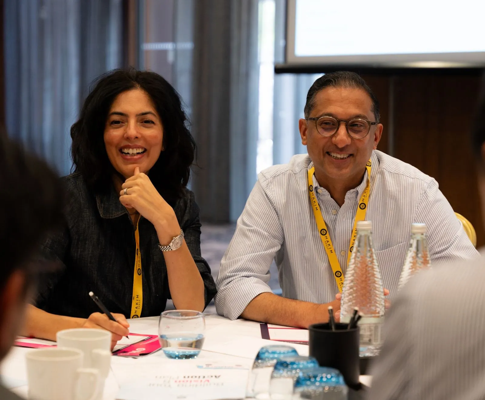 Two people wearing yellow lanyards sitting at a table, smiling and engaged in discussion with water bottles and cups on the table.