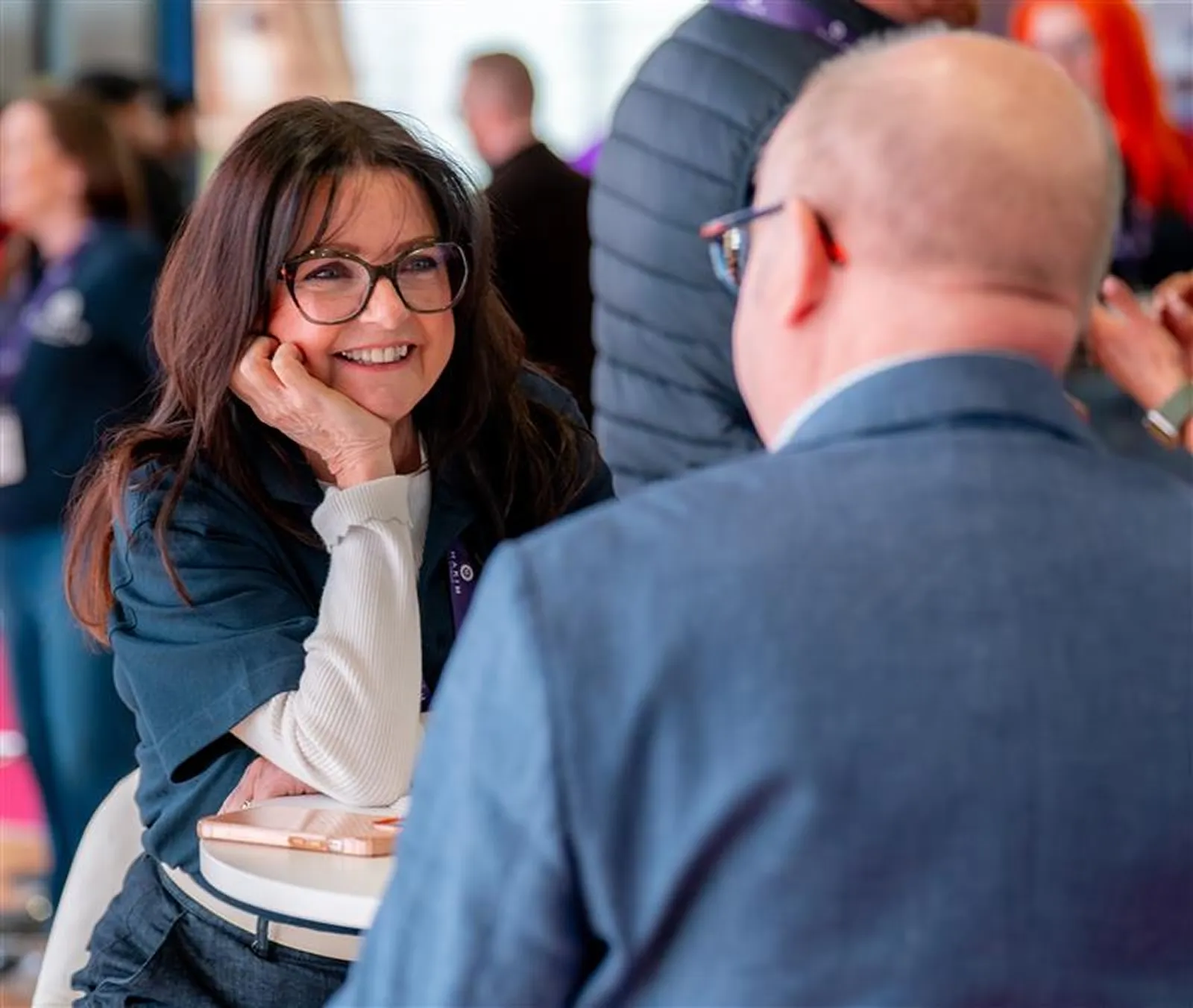 Smiling woman with glasses leaning on her hand engaged in conversation with a bald man in a suit at a table.