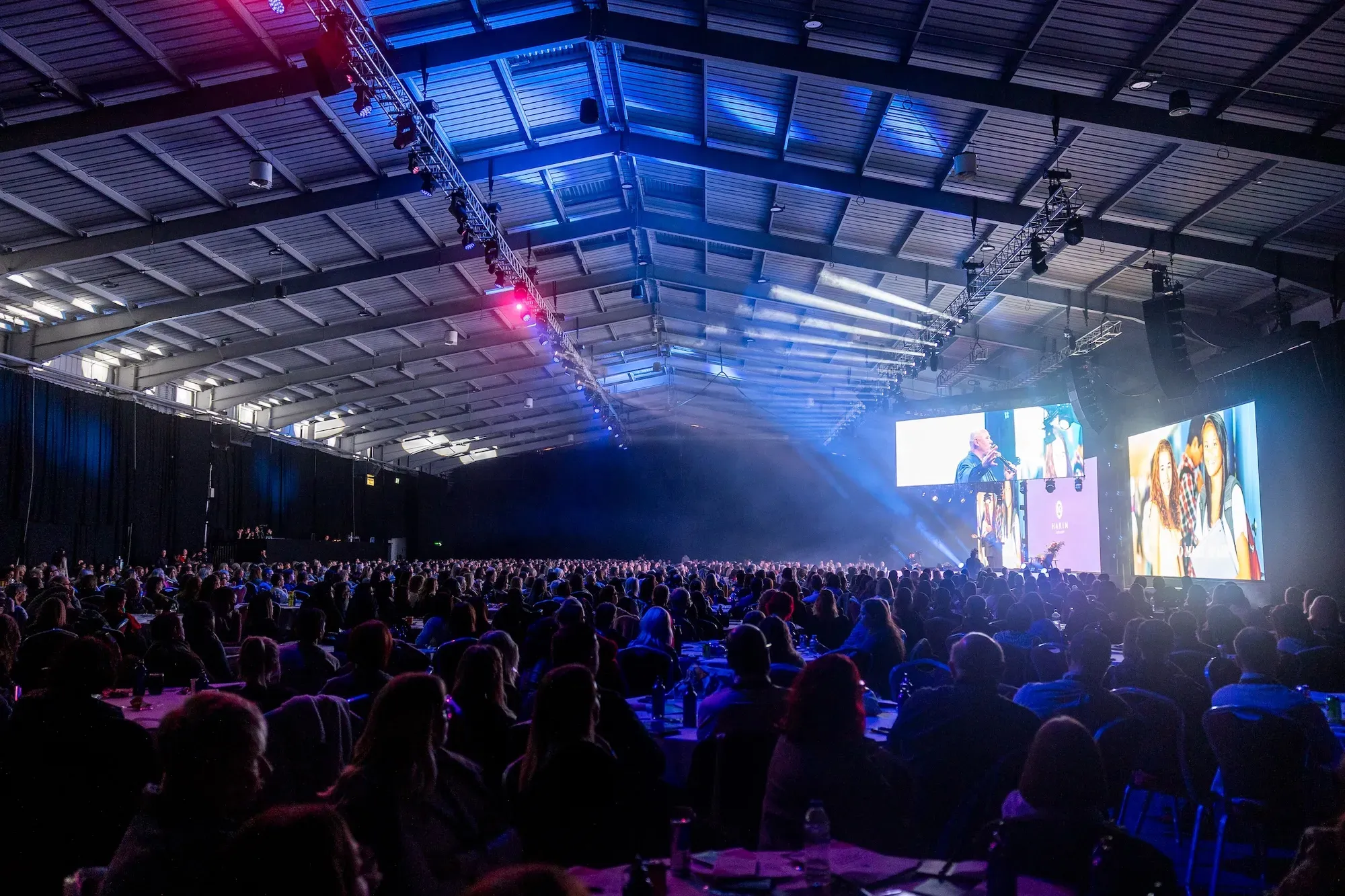 Large audience seated in a conference hall watching a speaker on stage with multiple large screens displaying images and presentations.