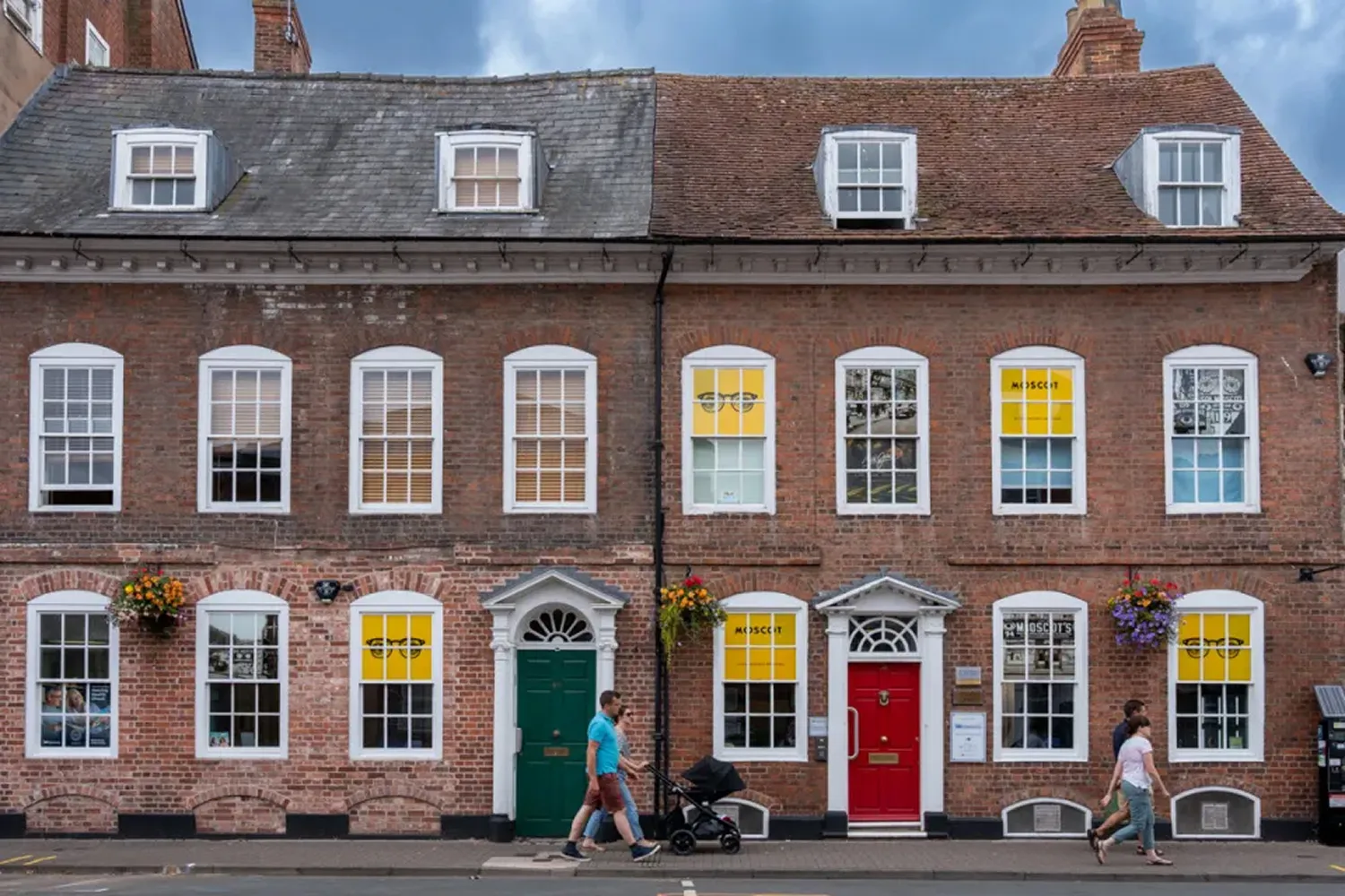 Historic brick building with white-framed windows, green and red doors, yellow window advertisements, and people walking past on the sidewalk.