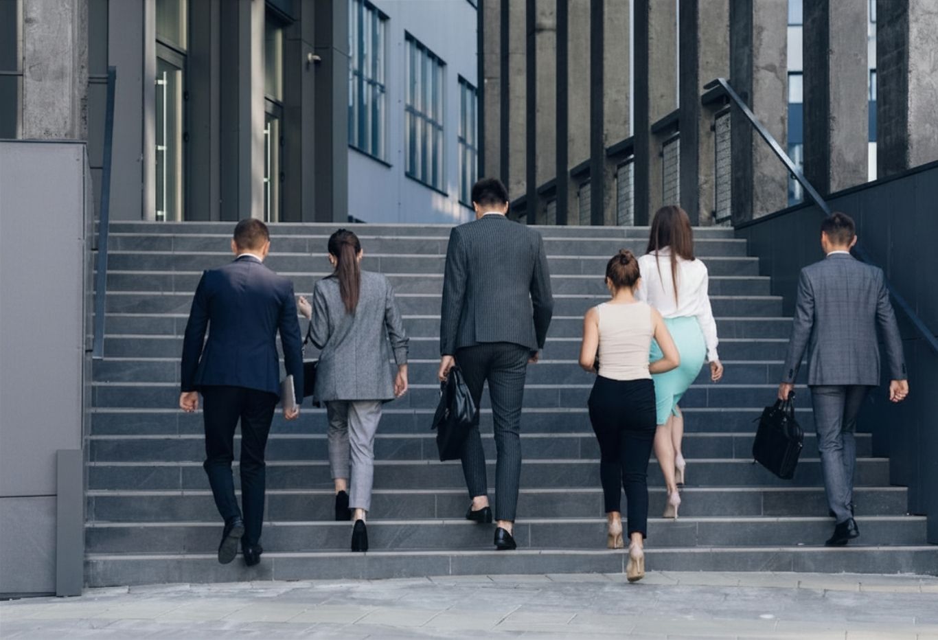Sechs Geschäftsleute in formeller Kleidung, drei Frauen und drei Männer, gehen eine Außentreppe in einem modernen Gebäudekomplex hinauf, zwei Männer tragen Aktentaschen.