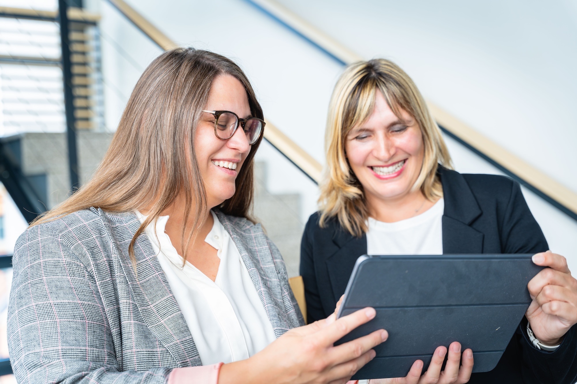 Zwei lächelnde Frauen, eine mit Brille und kariertem Blazer, die andere mit blondem Haar und dunklem Blazer, sehen gemeinsam auf ein Tablet.