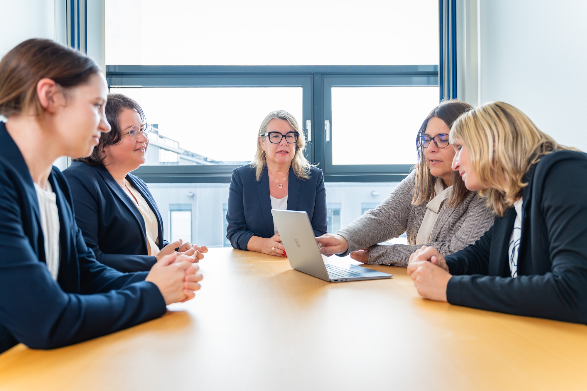 Fünf Frauen in Geschäftskleidung sitzen an einem hellen Konferenztisch, schauen auf einen Laptop, der in der Mitte steht, während eine Frau am Laptop auf den Bildschirm zeigt.