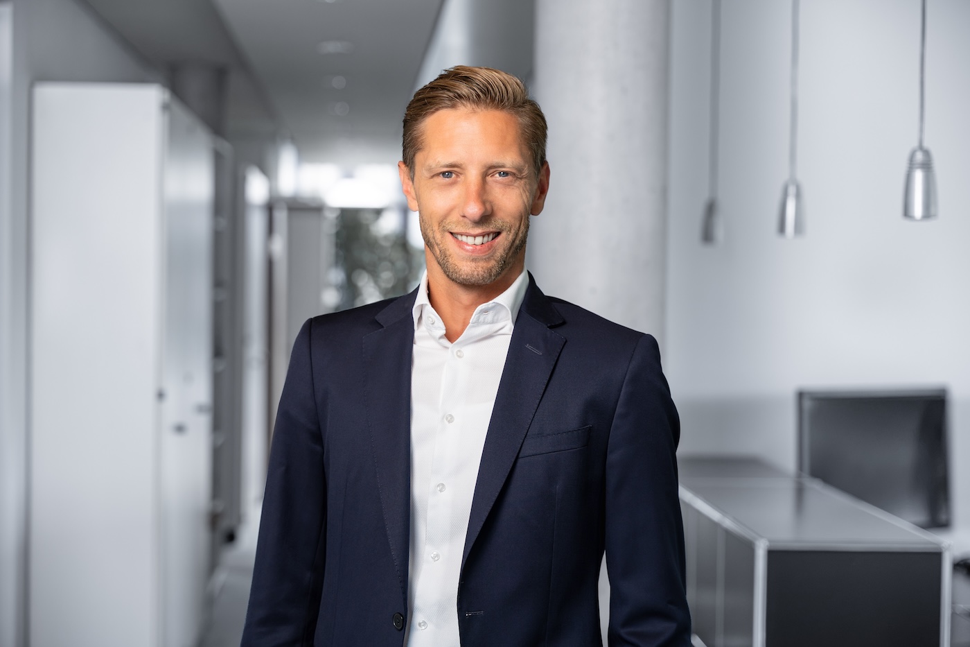 Porträt eines lächelnden Mannes mit blondem Haar, der einen dunklen Anzug und ein weißes Hemd in einem modernen Büro trägt.