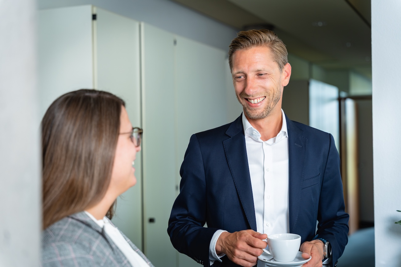 Ein lächelnder Mann im dunkelblauen Anzug hält eine weiße Tasse und einen Unterteller und unterhält sich mit einer Frau mit langen braunen Haaren und Brille in einem Büroflur.